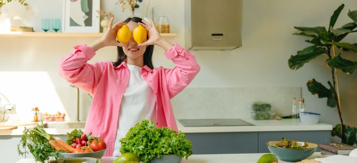 A woman playfully holding lemons over her eyes in a bright, lively kitchen setting filled with fresh vegetables.