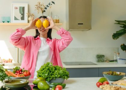 A woman playfully holding lemons over her eyes in a bright, lively kitchen setting filled with fresh vegetables.